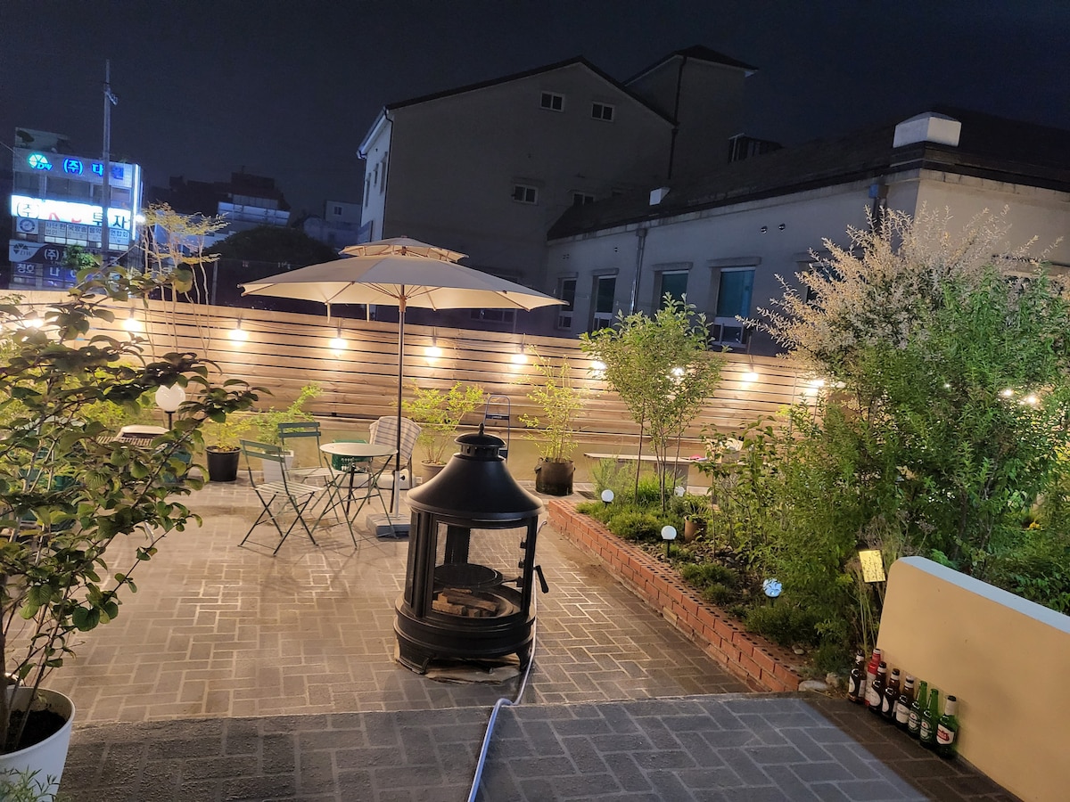 A well-lit garden area showcases outdoor seating with a table and chairs underneath an umbrella. A modern fire pit stands at the center, surrounded by greenery and plants. Soft lighting enhances the tranquil ambiance of the space during the night.