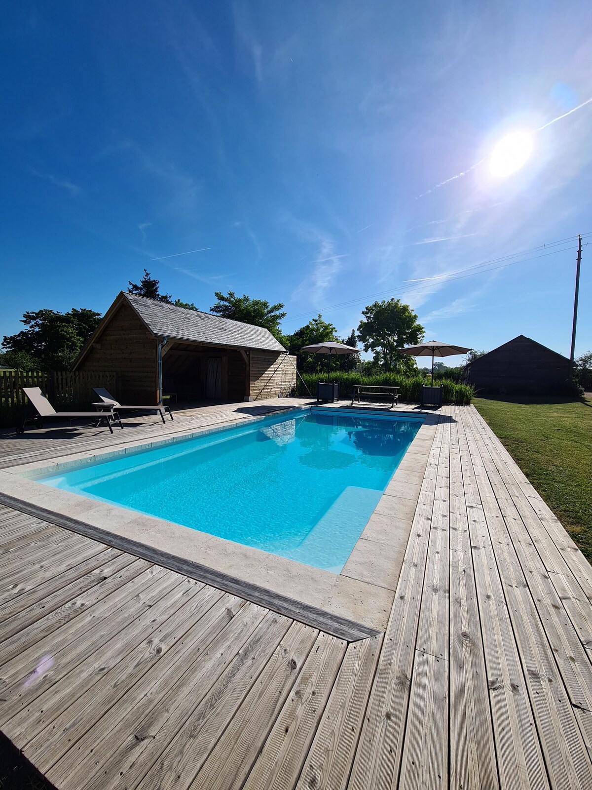 A heated swimming pool is surrounded by a wooden deck, with lounge chairs positioned for relaxation. A shaded area is visible in the background, providing options for outdoor seating. Clear sky and sunlight contribute to a bright atmosphere.