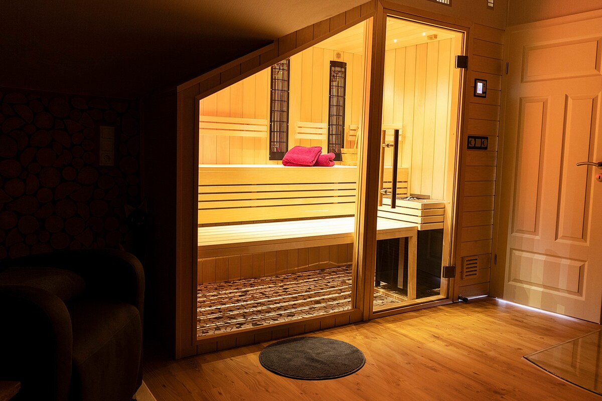 A modern sauna is featured with wooden benches and a plush towel resting on the backrest. Soft lighting illuminates the interior, enhancing the inviting atmosphere. The floor is finished in natural wood, and a black circular mat is positioned at the entrance.