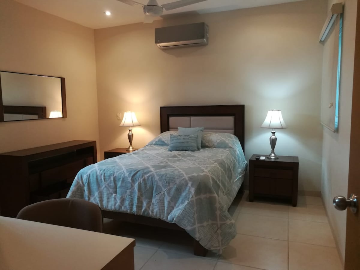 A well-appointed bedroom features a spacious bed with light blue bedding. Two bedside tables with lamps flank the bed, providing gentle illumination. A mirror reflects the room's soft tones, while a desk and chair are positioned for convenience. Natural light filters through the window.