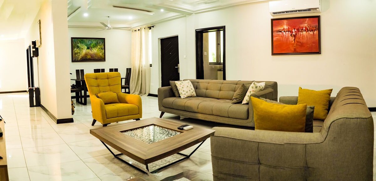A spacious living area features a grey sectional sofa and a bright yellow accent chair. A glass-top coffee table is centered on elegant tile flooring. Wall art adorns the walls, and large windows allow natural light to brighten the space.