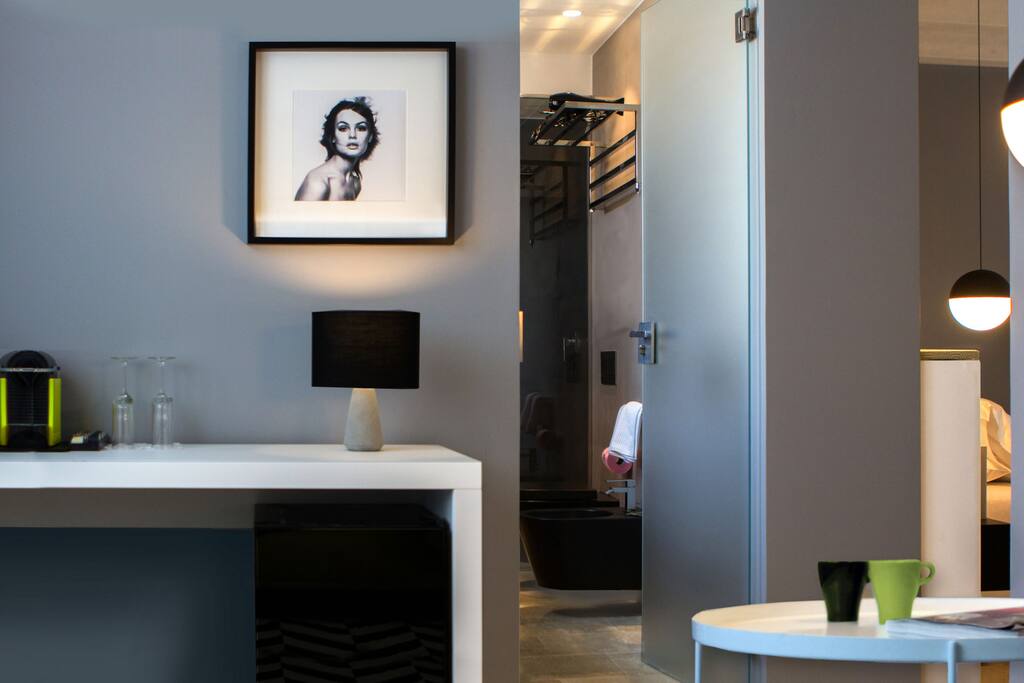 Al suo ingresso una spaziosa camera da letto total white che risalta tra le pareti e i dettagli grigi contornata da particolari lampade da terra e sedie intrecciate black and white che riprendono magicamente il tappeto del soggiorno accanto. Alla sinistra armadio a vista verde psichedelico disegnato e progettato su misura esclusivamente per CinqueSuites. Ampio bagno total black con elementi matt black che fanno da contrasto alle venature del legno che regge il lavabo dalle forme decise ma eleganti. Il soggiorno con il suo mood ever green, in cui la speciale lampada String Light di Flos crea il suo gioco di fili da una parete all’altra. Con balconcino che si affaccia sul suggestivo Ronco Purgatorio. L’intera struttura è pavimentata con tradizionale pietra di Modica siciliana.
