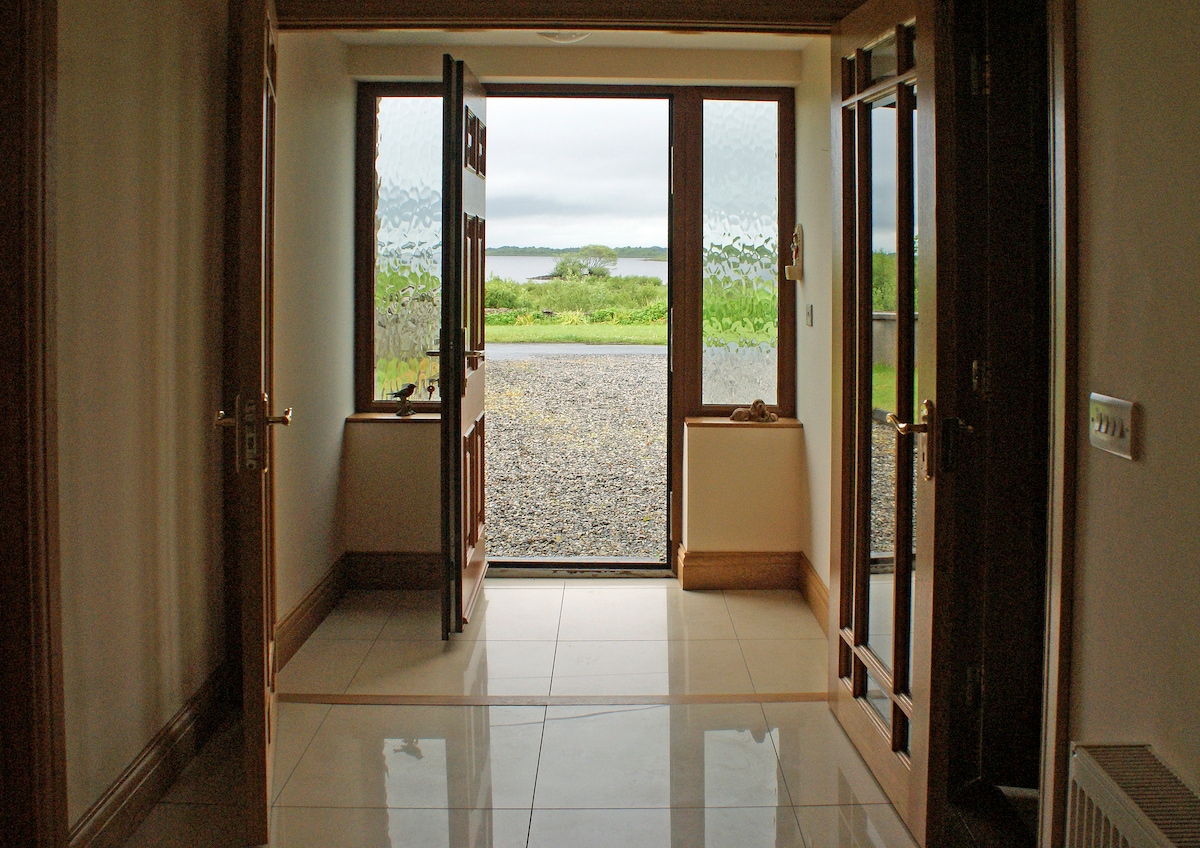 The entrance features large double doors made of wood, opening towards a scenic view of the lush green landscape and water beyond. A tiled floor reflects natural light, and the walls are neutrally painted, creating a welcoming space that connects indoors with the outdoors.
