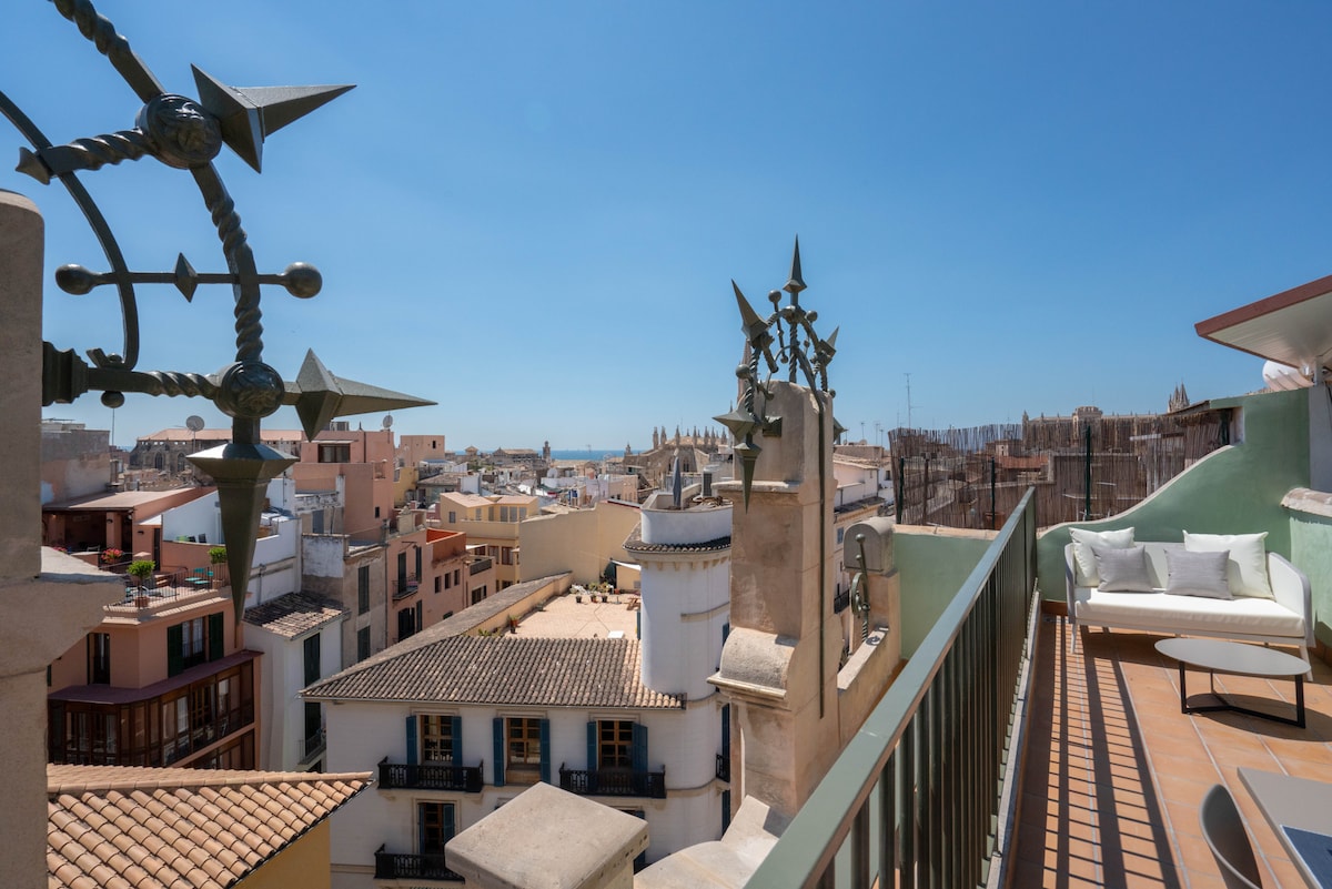 An outdoor terrace provides sweeping views of the old town of Palma and the sea. The space is furnished with lounge chairs and a small table, while architectural details, such as wrought iron features, adorn the surrounding edges. Clear blue skies provide a bright backdrop.