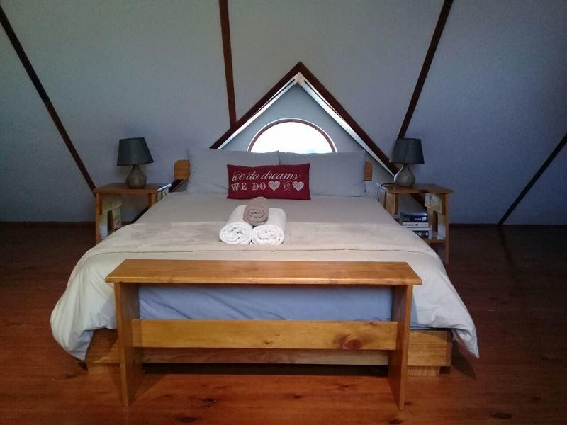 A spacious mezzanine bedroom features a wooden bed with neutral bedding and decorative pillows. Two bedside tables with lamps are positioned on either side. A circular window allows natural light to enter, complementing the wooden flooring and sloped ceiling.