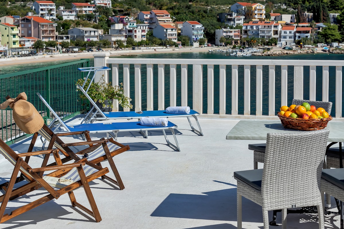 A shared terrace is visible, featuring two lounge chairs, a wooden rocking chair, and a dining table set. A bowl of oranges is placed on the table. The serene view of the water and buildings in the background adds to the outdoor space's appeal.