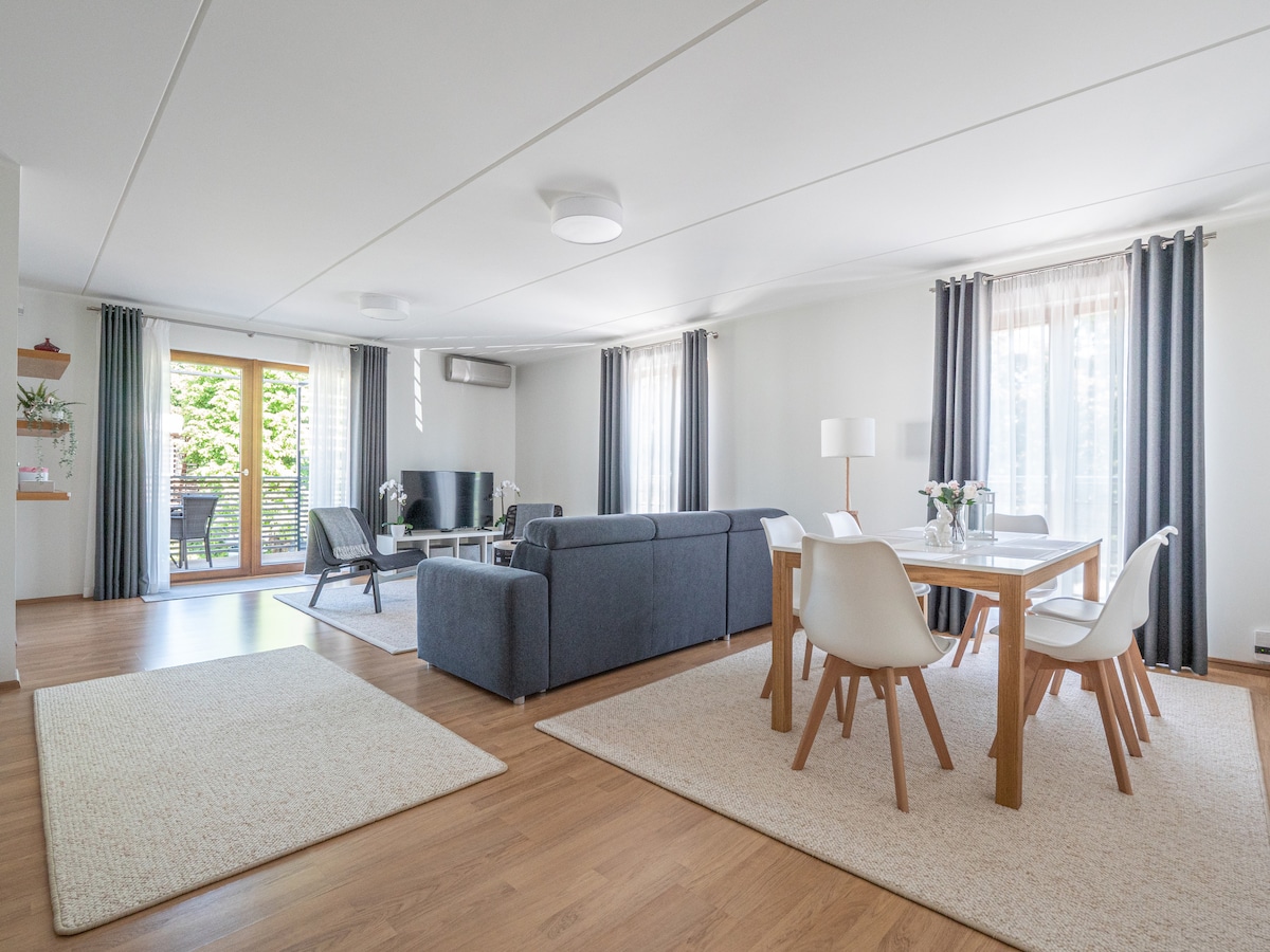 An open plan living area is presented with a comfortable sofa and a dining table with six chairs. Natural light filters through large windows covered with light curtains, creating a bright space. A TV is positioned to one side, with a balcony visible through the sliding doors.