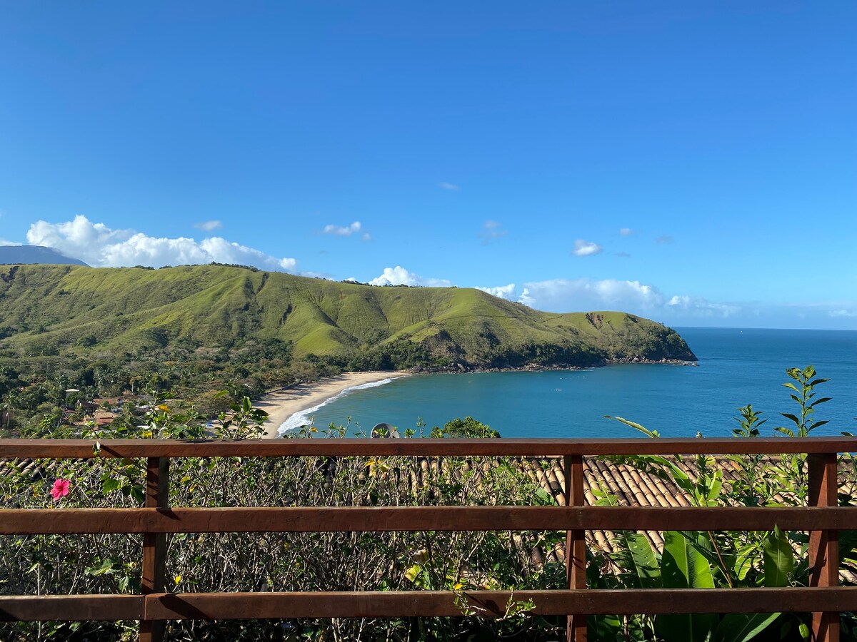 A scenic view of a tranquil beach and lush green hills is captured from a wooden deck. The coastline curves gently into the distance, while the clear blue water reflects the bright sky adorned with a few white clouds.