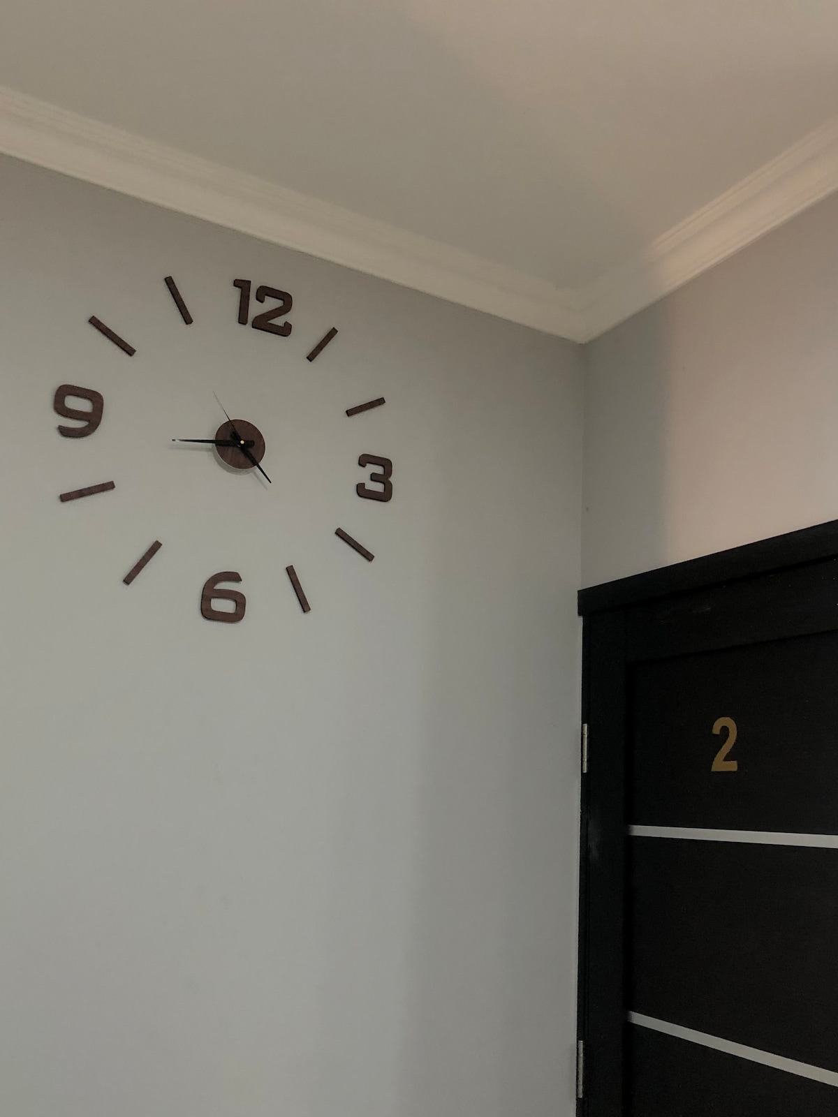 A large wall clock is mounted above a doorway, featuring bold, easy-to-read numbers at each hour mark. The clock hands are positioned at approximately 10:10. The walls are painted in a soft gray, complementing the dark door frame labeled with the number '2'.