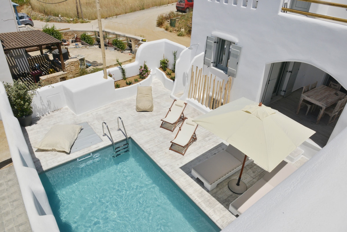 An outdoor area features a private pool surrounded by a spacious terrace. Sun loungers and a large umbrella are positioned for relaxation. Whitewashed walls complement the natural surroundings, with patches of greenery adding a fresh touch. The setting invites leisurely moments under the sun.