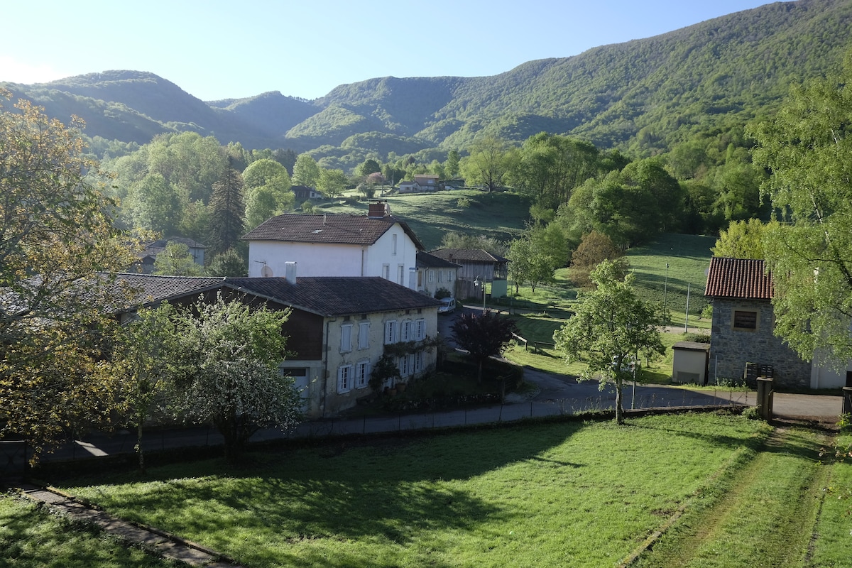 A rural landscape is showcased, featuring a cluster of charming buildings nestled among lush green hills. Vibrant trees and open fields are visible, with the distant mountains providing a serene backdrop. The early morning light casts a gentle glow across the scene.