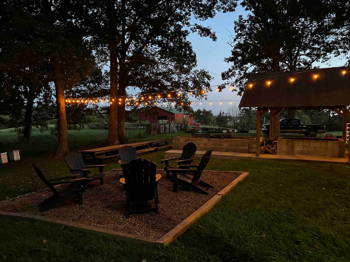 A cozy outdoor gathering area features several Adirondack chairs arranged around a gravel fire pit, illuminated by string lights overhead. A rustic shelter is visible, alongside a spacious lawn and gently rolling hills in the background.