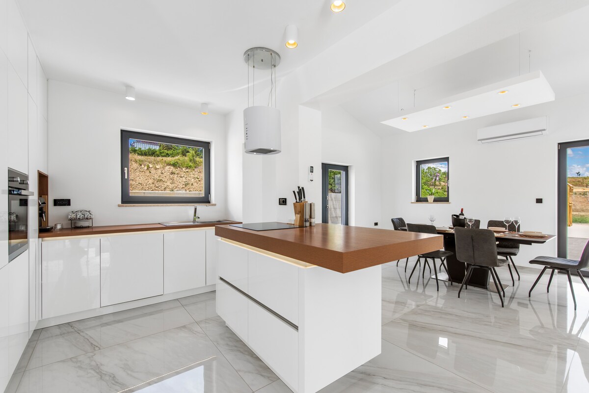 A modern kitchen features sleek white cabinetry, an integrated oven, and a prominent island with a wooden countertop. Natural light floods the space through large windows, which offer views of the surrounding landscape. The adjacent dining area is furnished with a stylish table and comfortable chairs.