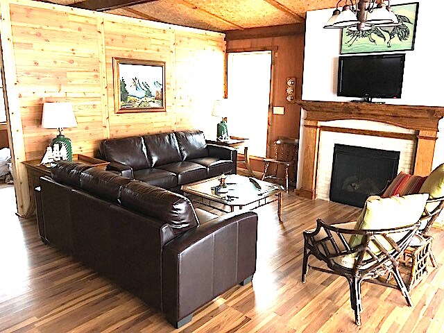 A warm living area features a dark leather sofa facing a glass coffee table. Two lamps provide illumination beside the windows, and a fireplace is set into the wall below a mounted television. A decorative painting is displayed above the mantel.
