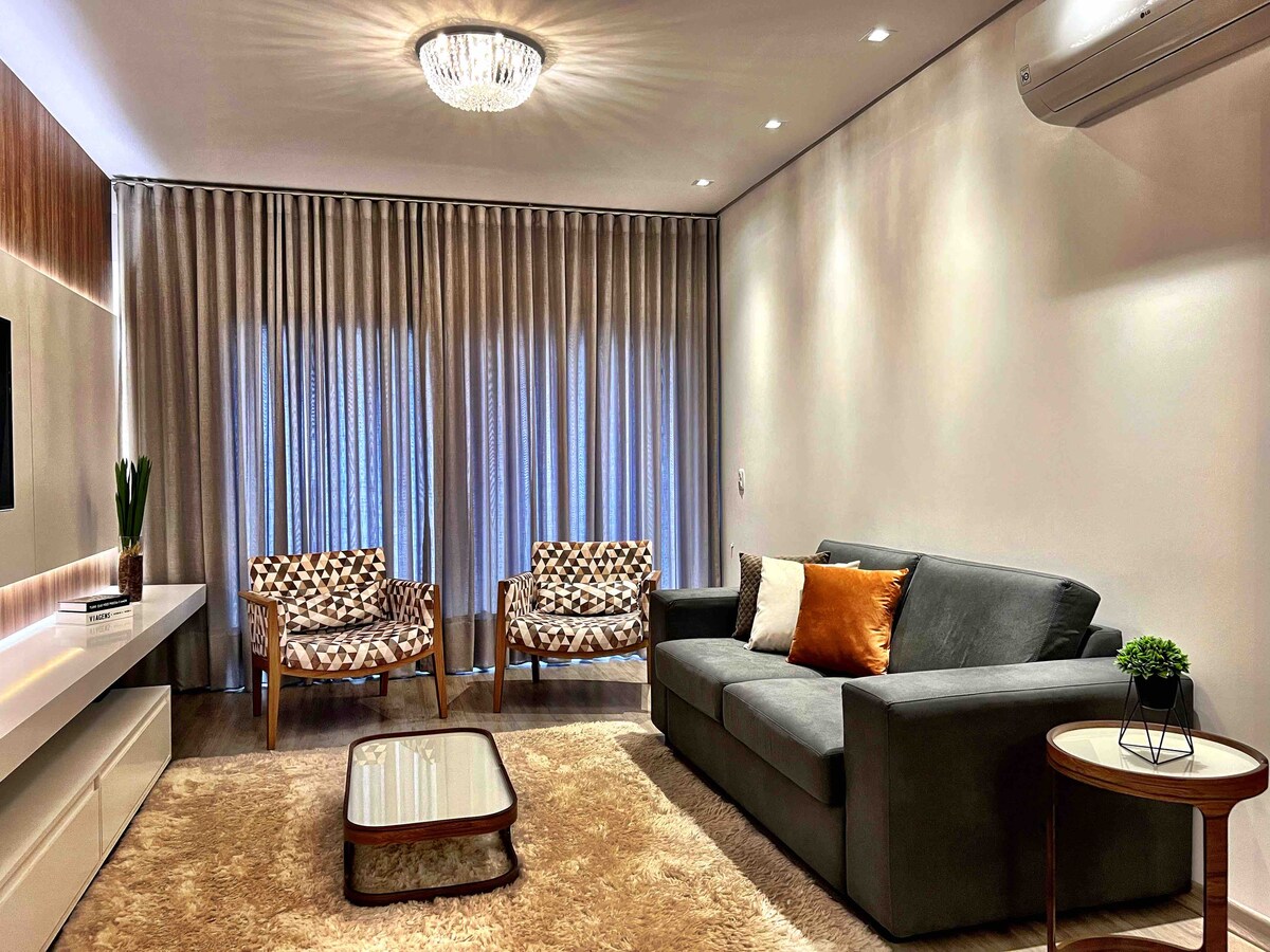 A modern living area is presented with a gray sofa complemented by an orange throw pillow. Two patterned chairs are positioned opposite the sofa. Large windows are dressed with light curtains, allowing natural light to fill the space, while a small round table and a plant add a touch of decor.