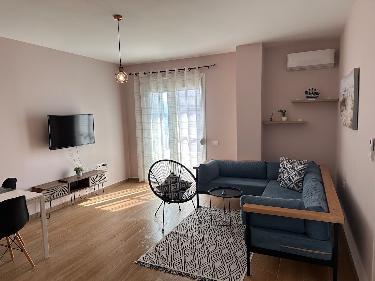 A living area features a comfortable blue sofa paired with a black accent table. A television is mounted on the wall, while large glass doors lead to a balcony. Natural light fills the space through sheer curtains, complemented by a patterned area rug on the tiled floor.