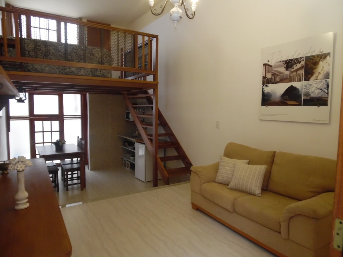 A spacious living area features a light-coloured sofa with decorative cushions. A wooden dining table is visible nearby, accompanied by a set of chairs. A staircase leads to a lofted area, while natural light filters through large windows.