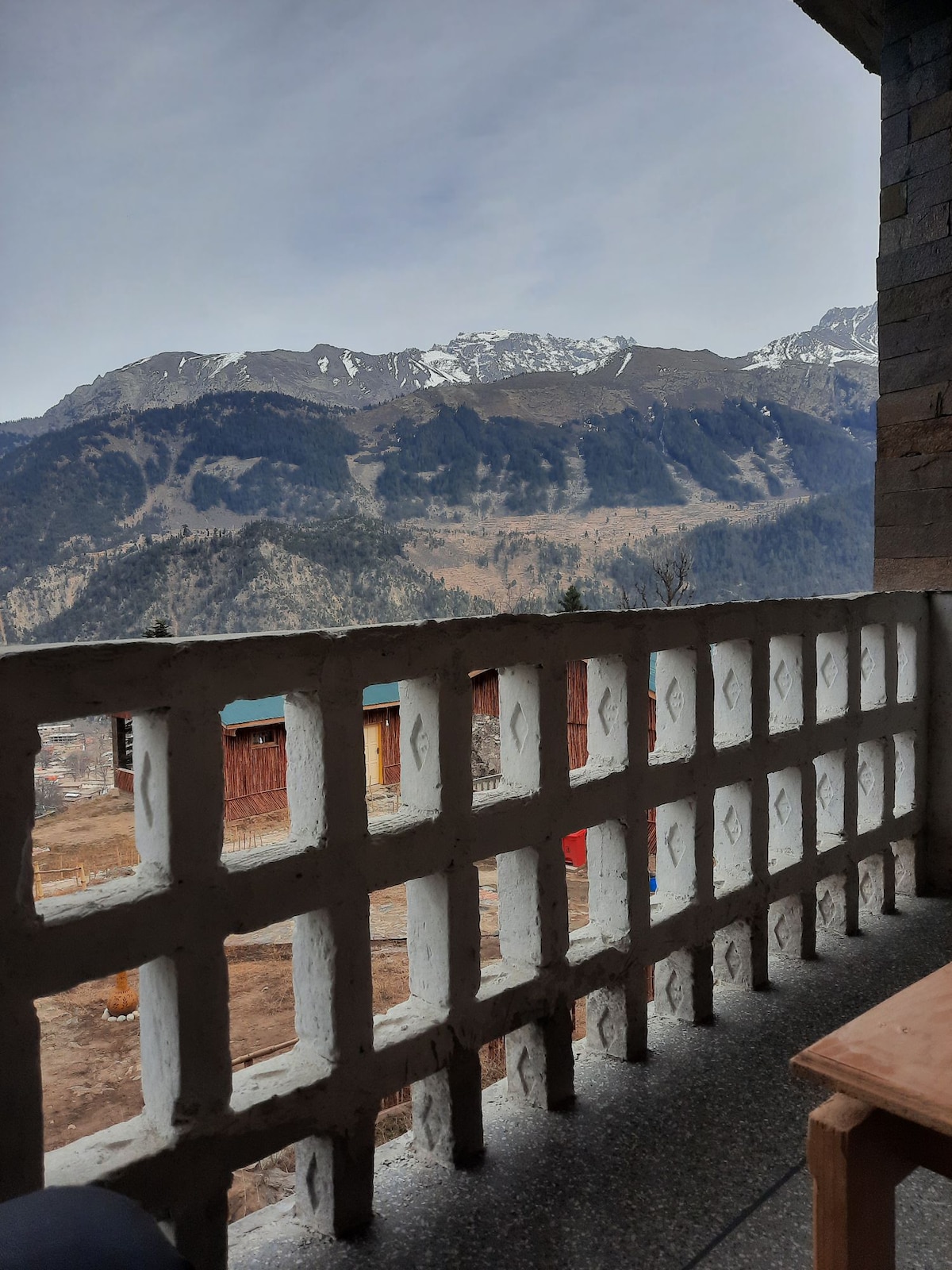 A view from the terrace showcases a mountain landscape, with snow-capped peaks rising in the background. The foreground features a stone railing and a wooden table placed on a stone floor, creating a serene setup to enjoy the surrounding natural beauty.