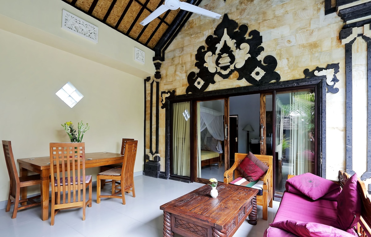 The living area showcases a wooden dining table with six chairs, complemented by a cozy seating area with burgundy cushions. Large glass doors invite natural light, revealing a connection to the outside terrace. The room features traditional Balinese architectural details, enhancing its aesthetic appeal.