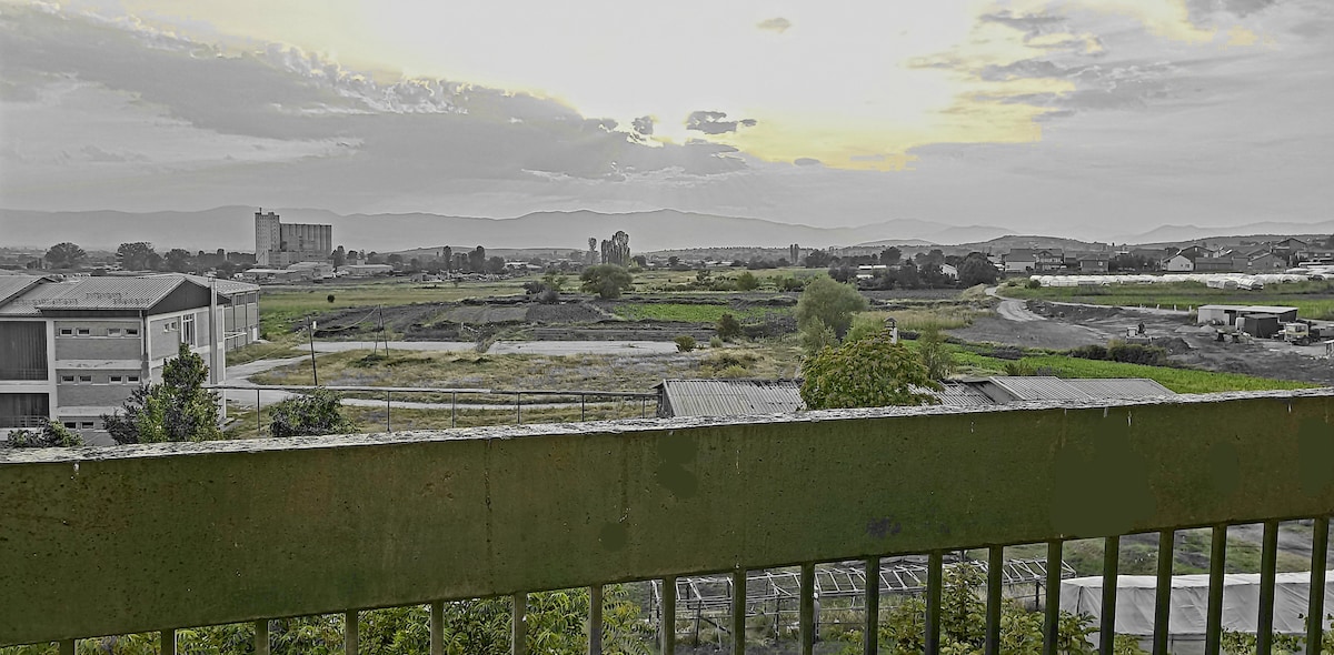 A panoramic view captures an expansive landscape featuring fields and distant mountains under a soft sunset. Industrial buildings can be seen in the background, with a blend of lush greenery and open spaces stretching across the horizon.