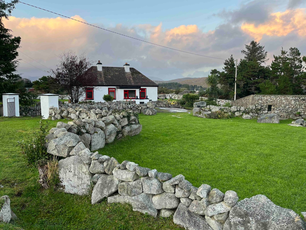 A traditional cottage is set against a backdrop of rolling hills and vibrant sky. The surrounding area features well-maintained grass and low stone walls. Native trees frame the property, enhancing the rural ambiance of this peaceful setting.
