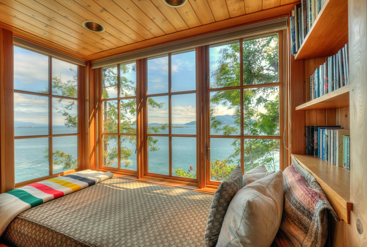 A cozy window seat is surrounded by large windows, offering panoramic views of the water and trees outside. Soft throw pillows and a blanket are arranged on the natural fiber mattress, creating a relaxing spot to enjoy the scenic surroundings.