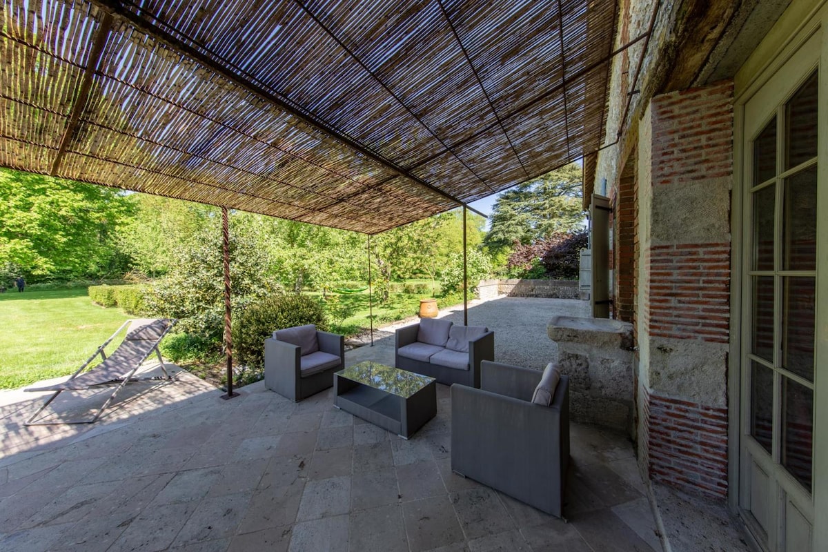 A shaded outdoor seating area is presented under a thatched roof, featuring a glass table surrounded by comfortable grey armchairs. The lush greenery of the garden is visible in the background, enhancing the serene ambiance of the space.