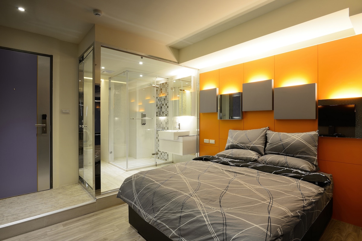A modern bedroom features a spacious bed with gray bedding, complemented by orange accent walls. The ensuite bathroom is visible through glass panels, showcasing stylish tile work and contemporary fixtures. Soft lighting enhances the overall ambiance, while a sitting area provides additional comfort.