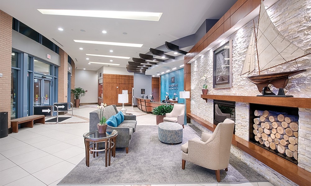 The lobby area is designed with modern furnishings, including two armchairs and a cream sofa. A coffee table sits on a soft rug, complemented by a decorative plant. A stone fireplace is adorned with a nautical sculpture, enhancing the inviting ambiance.