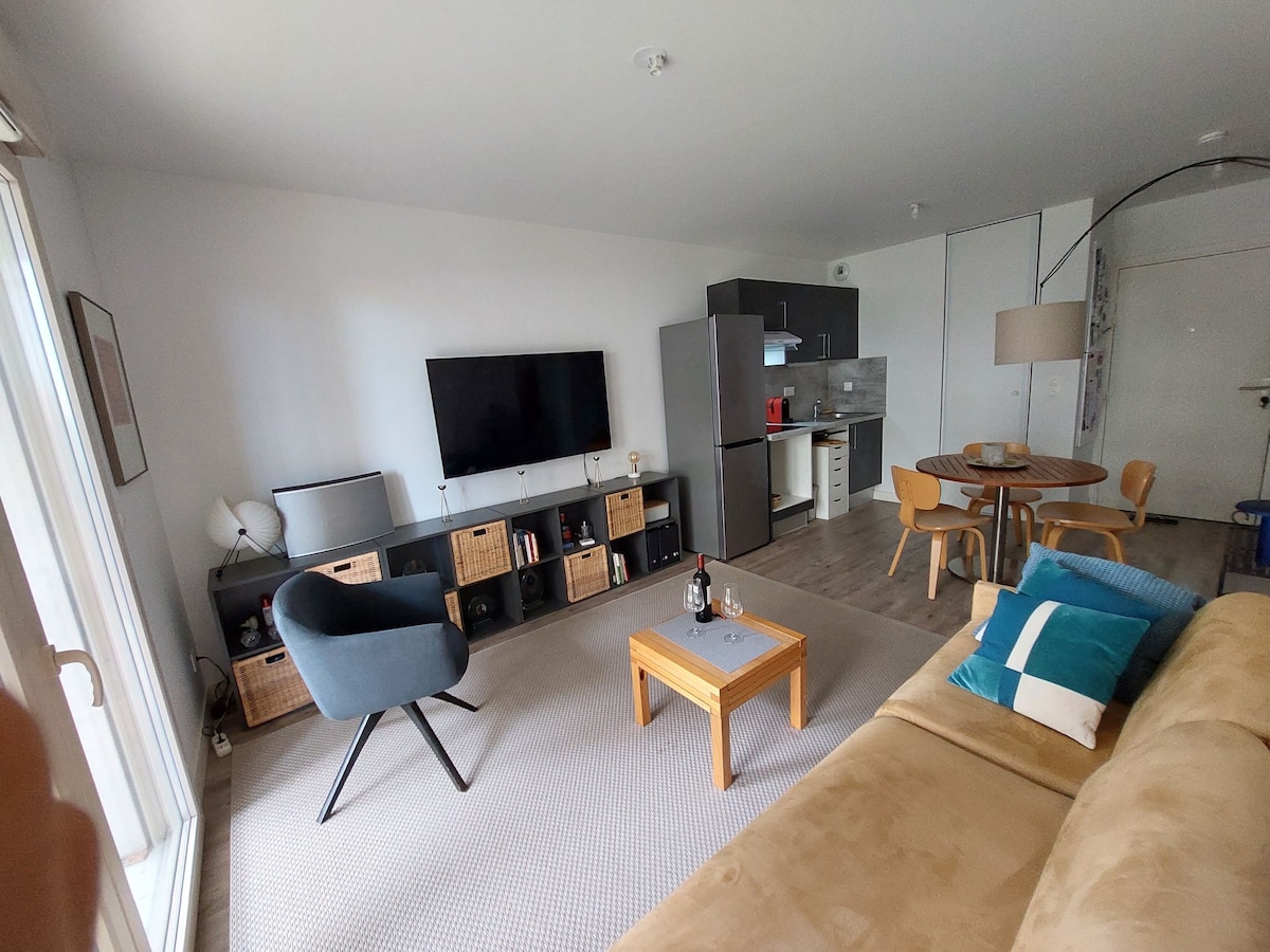 A spacious living area is presented, featuring a soft brown sofa and a circular dining table surrounded by chairs. A large television is mounted on a wall unit, complemented by storage baskets. Natural light is welcomed through glass doors leading to a balcony.