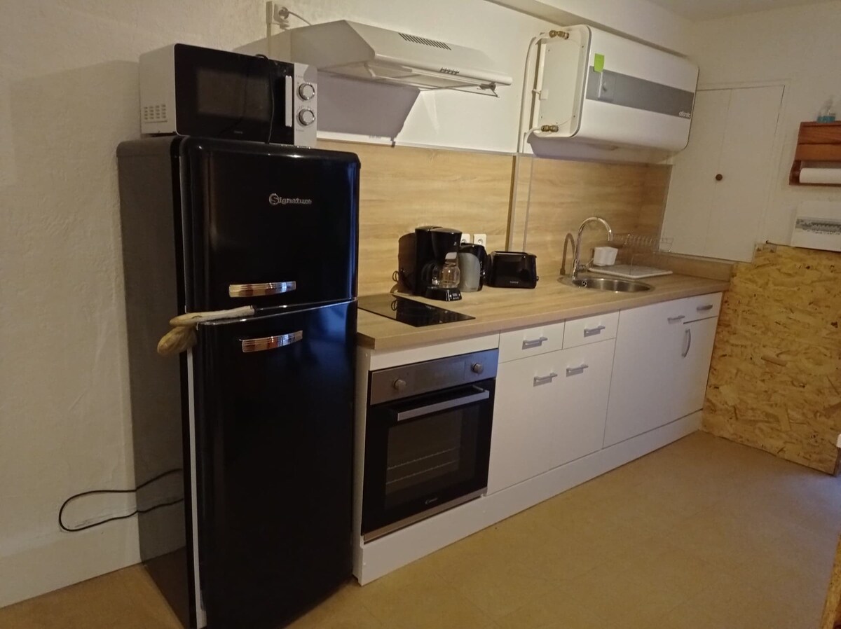 A modern kitchen area is equipped with essential appliances including a black refrigerator, a microwave, and a coffee maker. Counter space features a stainless steel sink and an oven, while light-colored cabinetry provides functional storage. The overall layout maintains a clean and organized appearance.