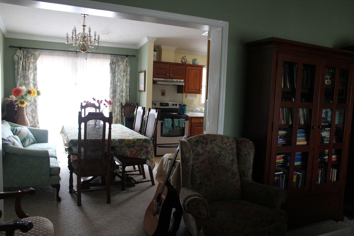 A shared dining and living space features a large table set with floral accents. A comfortable armchair and a shelving unit filled with books are visible, alongside a well-equipped kitchen. Natural light flows through the window, enhancing the inviting atmosphere of the room.