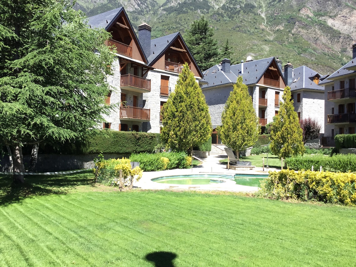 A landscaped garden area is visible, featuring well-maintained lawns and neatly trimmed hedges. Several stone buildings with wooden balconies surround the space. A circular pool is situated at the center, with paths lined by greenery leading to the pool area.