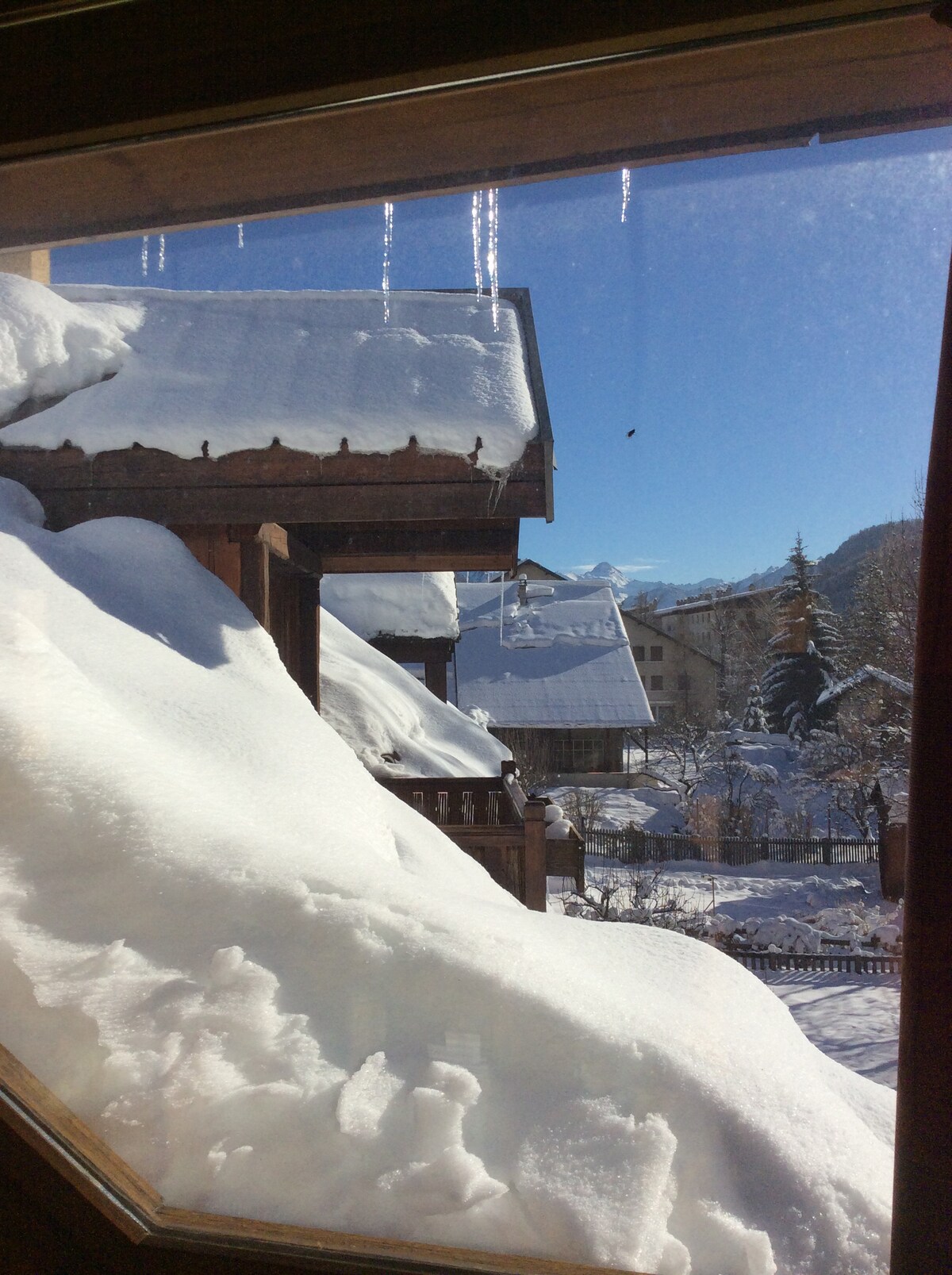Snow-covered rooftops are visible from the window, with a clear blue sky above. Icicles hang from the eaves, and a winter landscape is seen beyond, featuring trees dusted with snow and charming village architecture.
