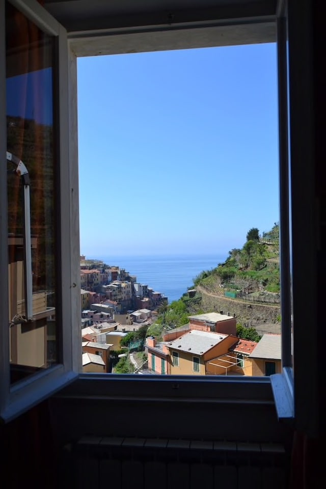 A window frame opens to a clear view of the coastline and colorful buildings of Manarola. The azure sea can be seen blending into the horizon under a bright blue sky, while the terraced landscape adds depth to the scene.