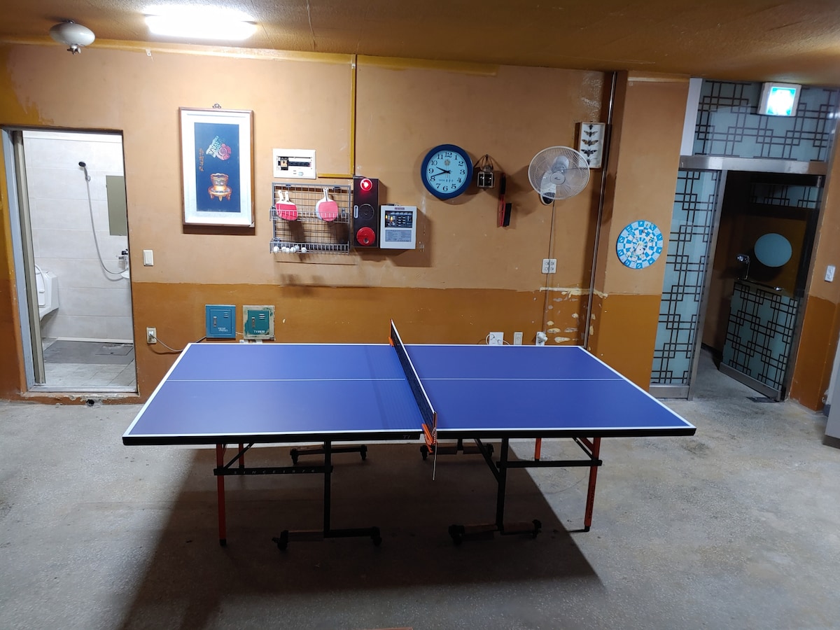 A table tennis table occupies the center of a spacious room, with a smooth blue surface and red net. Walls are adorned with decorative clocks and artwork, while a restroom is visible in the background. A fan is mounted on the wall for added comfort.