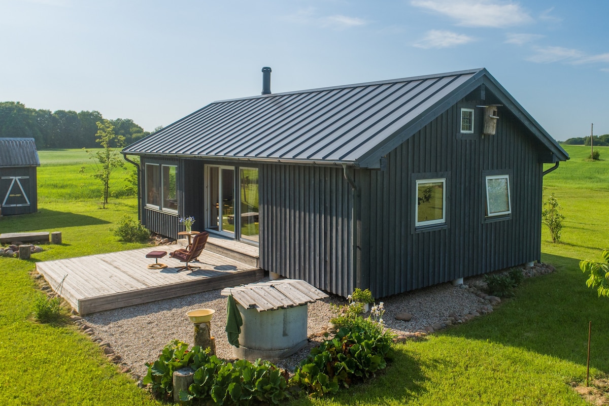 A modern cottage features a wooden exterior with a metal roof, surrounded by green fields. A wooden deck is located at the front, along with comfortable seating and a small outdoor table, inviting relaxation in a natural setting.
