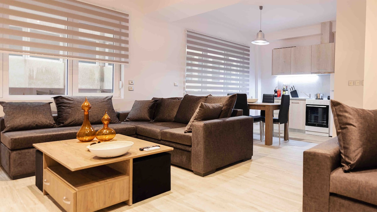 A spacious living area is showcased, featuring a sectional sofa adorned with cushions and a wooden coffee table. Natural light filters through dual-layered window shades, illuminating the open layout that includes a dining table and a modern kitchen setup.