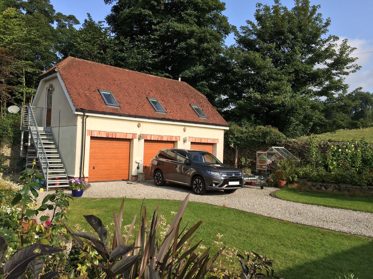 The exterior of the detached garage features a two-story design with a red tiled roof and large garage doors. A gravel driveway leads to the entrance, while an outdoor staircase provides access to the apartment above. Lush greenery surrounds the property, enhancing the rural setting.