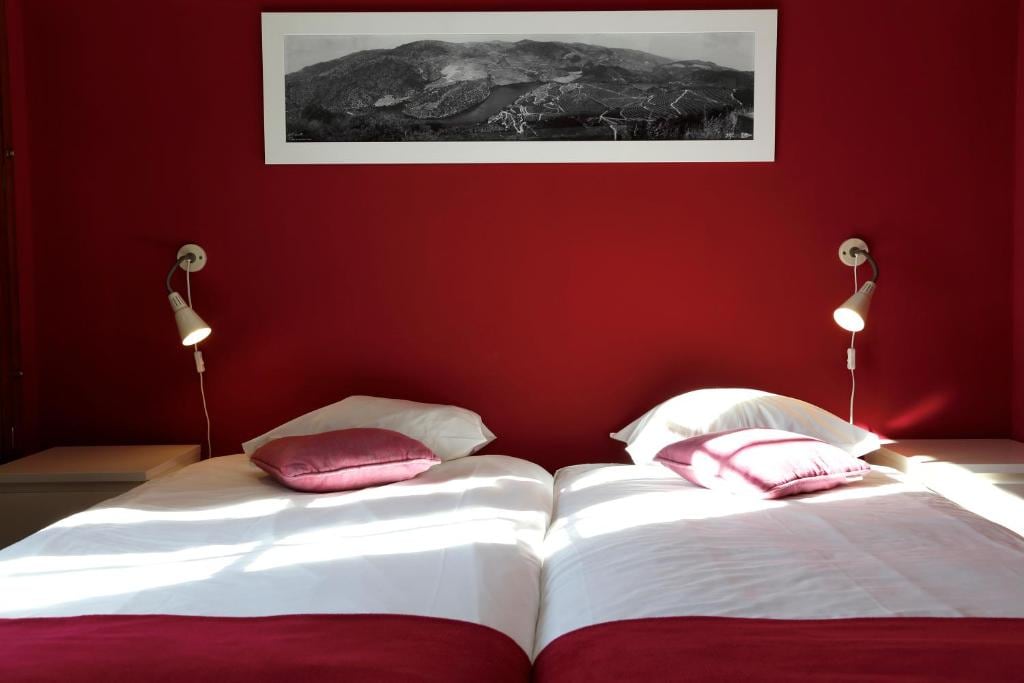 A vibrant red wall frames a well-lit sleeping area featuring two spacious beds with white linens. Each bed is complemented by a decorative cushion, and reading lamps are mounted on the wall above. A panoramic black-and-white photograph hangs above, adding visual interest.