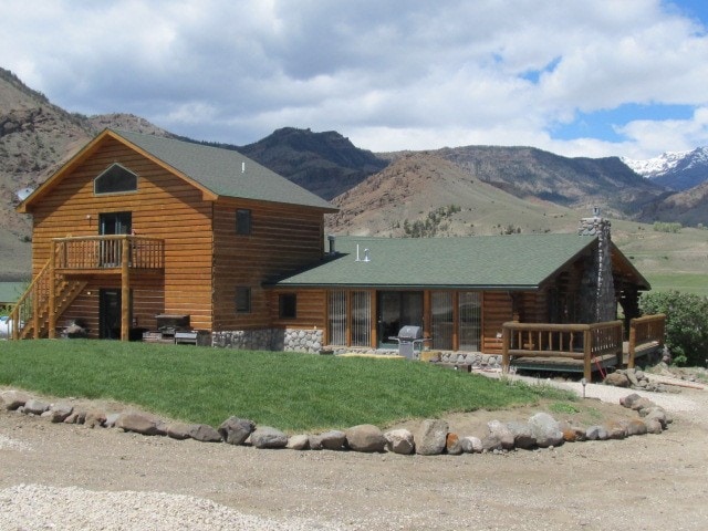 A spacious wooden cabin with a two-story design is situated against a mountainous backdrop. The exterior features a green roof, stone accents, and a wrap-around porch. Large windows enhance natural light, while a grass area and gravel pathway provide a welcoming entry.