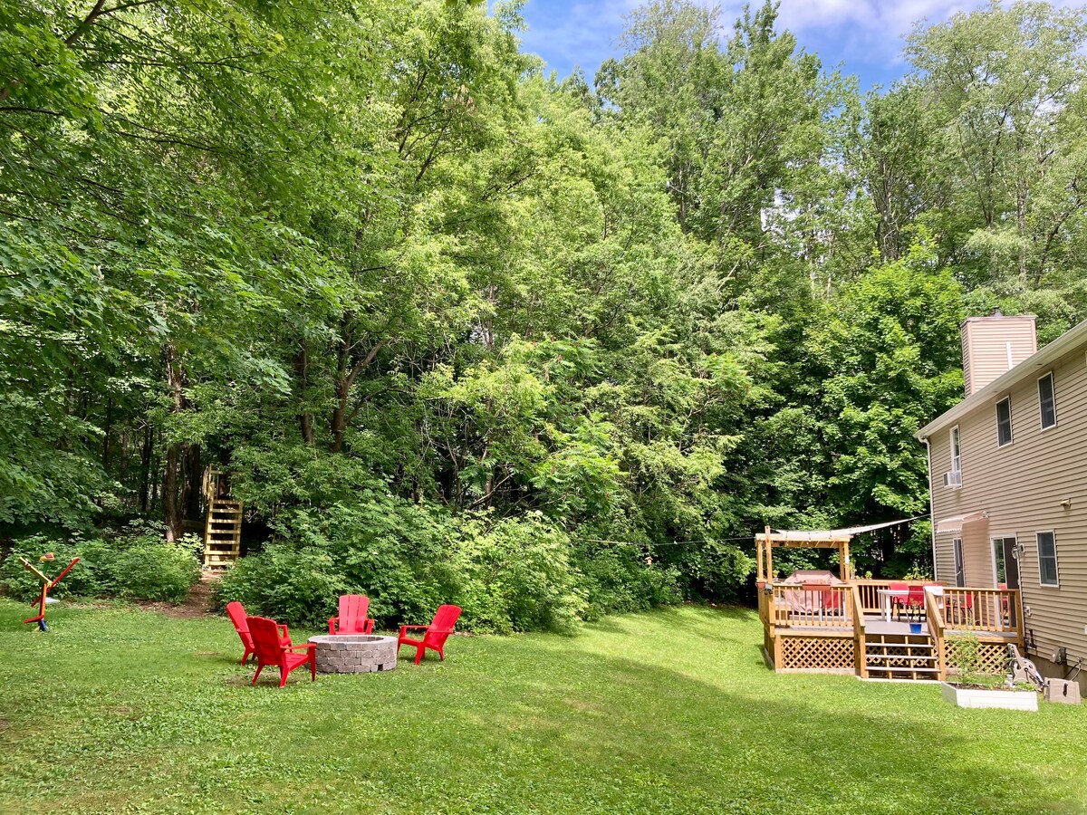 A grassy yard is framed by tall trees, creating a serene natural setting. A circular stone fire pit is surrounded by bright red chairs, offering a cozy spot for gatherings. A spacious deck with seating is visible adjacent to the house, providing an ideal area for relaxation.