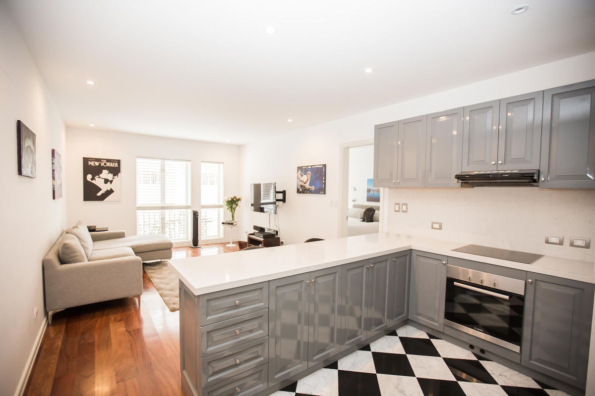 An open kitchen area with gray cabinetry showcases a modern design. A black and white checkered floor leads into a spacious living area featuring a light-colored sofa. Natural light fills the space through large windows, enhancing the bright, clean atmosphere.
