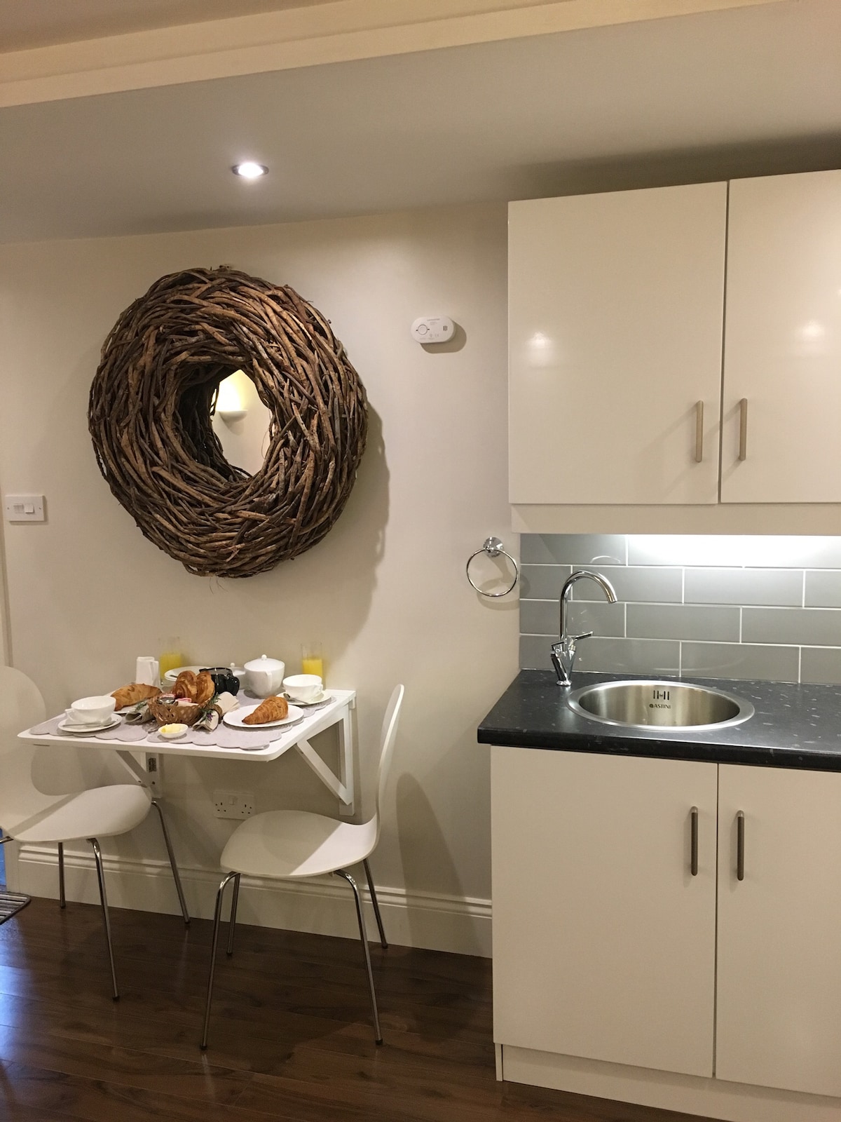 A small dining area is displayed with a round white table and two white chairs. A tray with breakfast items, including pastries and drinks, is set on the table. A rustic wooden mirror enhances the wall above the table. Kitchen cabinetry is arranged beside the sink.