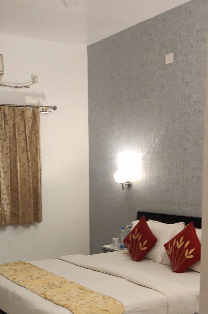 A comfortable sleeping area is highlighted by a spacious bed with decorative red pillows. A gold-accented bed runner adds a touch of elegance. Natural light filters through a gold-toned window curtain, while a light fixture illuminates the textured wall behind.
