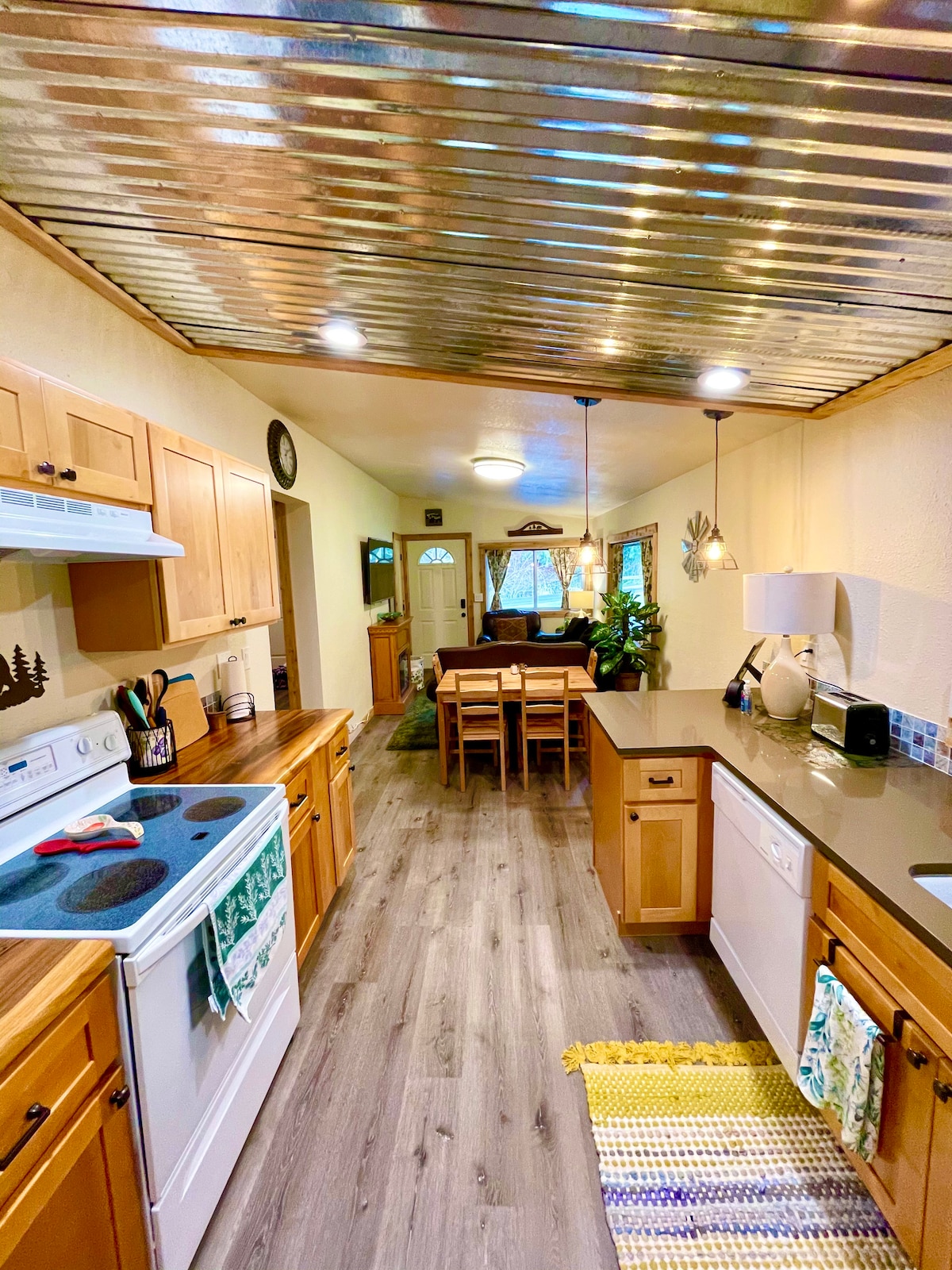 The kitchen is equipped with wooden cabinets and modern appliances, including a stove and dishwasher. A countertop provides ample space for meal preparation. A dining area with a wooden table and chairs can be seen in the background, illuminated by natural light from nearby windows.