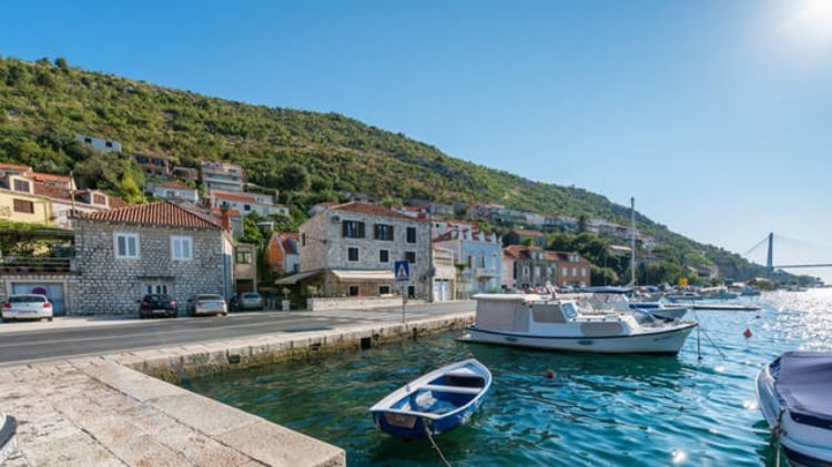 A serene waterway is lined with stone buildings and boats anchored at the shore. Lush greenery blankets the hillside in the background, while a bright sky reflects on the calm water surface. The scene is complemented by a distant bridge, enhancing the picturesque setting.