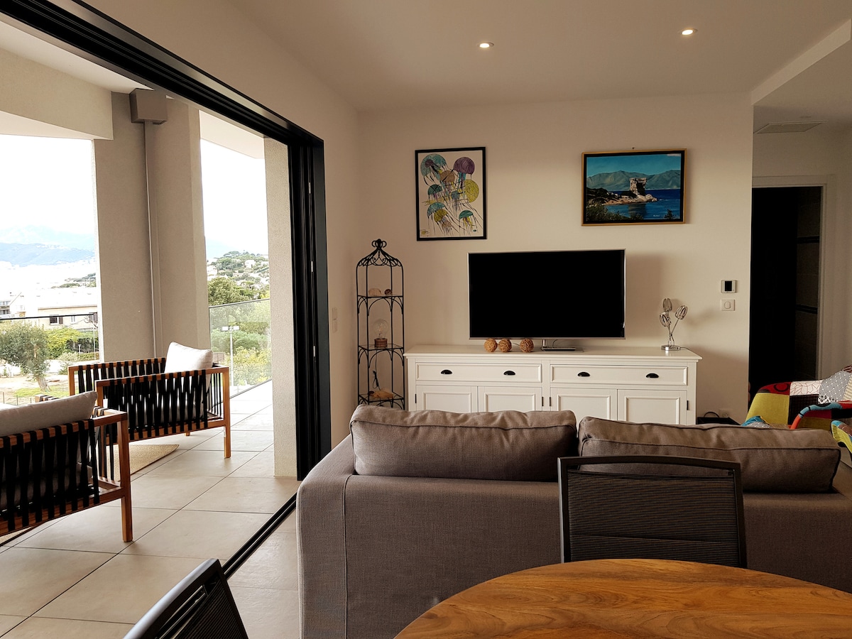 A spacious living area is shown with large glass doors leading to a terrace. A contemporary sofa faces a flat-screen TV, while two armchairs are situated nearby. Decorative artwork is displayed on the walls, and a circular dining table is positioned in the foreground.
