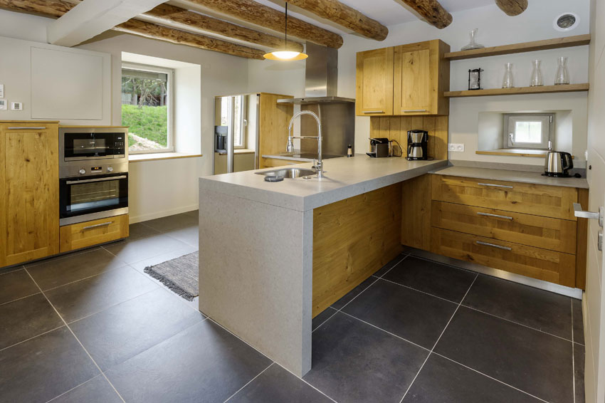 The kitchen features modern wooden cabinetry paired with sleek stone countertops. Stainless steel appliances, including an oven and refrigerator, are integrated into the design. A large window allows natural light to illuminate the space, highlighting the rustic wooden beams overhead.