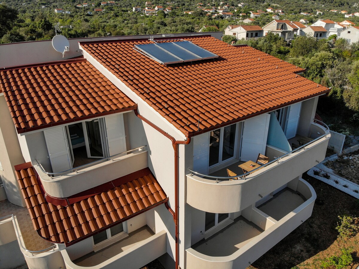 An aerial view of a two-story apartment complex is displayed, showcasing a terracotta-tiled roof and large glass windows. Balconies with slight overhangs are featured on each level, overlooking the surrounding greenery and residential areas.